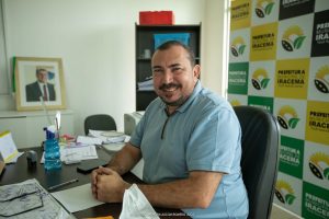 Jairo Ribeiro sorri sentado em seu gabinete na Prefeitura com foto de Jucá no fundo